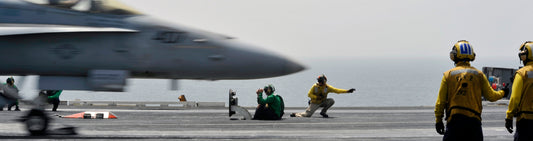 Close-up image of an aircraft carrier deck, highlighting a jet in motion and the essential roles of the crew. The scene depicts the precision and teamwork required for successful aircraft carrier operations.